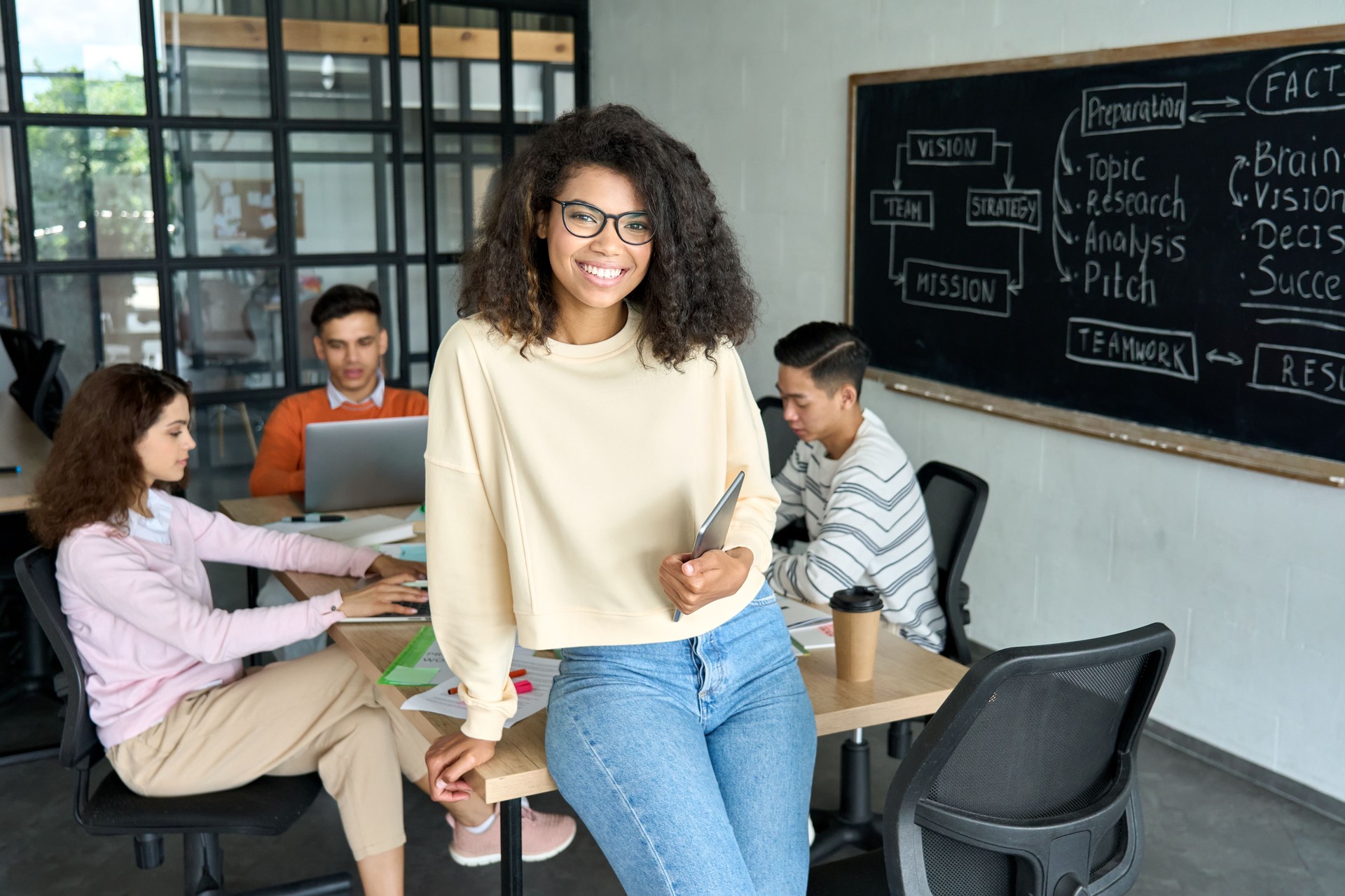 Young happy African American woman team leader sitting on desk holding tablet.